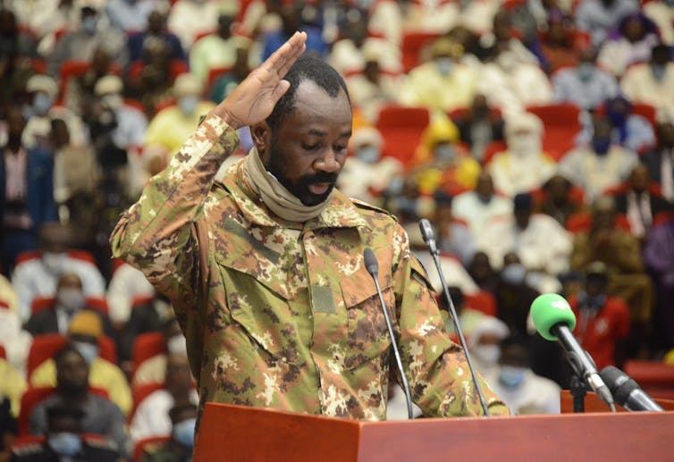 Goita, a middle-aged Black man, stands at a lectern in fatigues with his hand raised, with a crowd in foreground