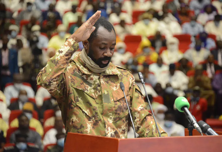 Goita, a middle-aged Black man, stands at a lectern in fatigues with his hand raised, with a crowd in foreground