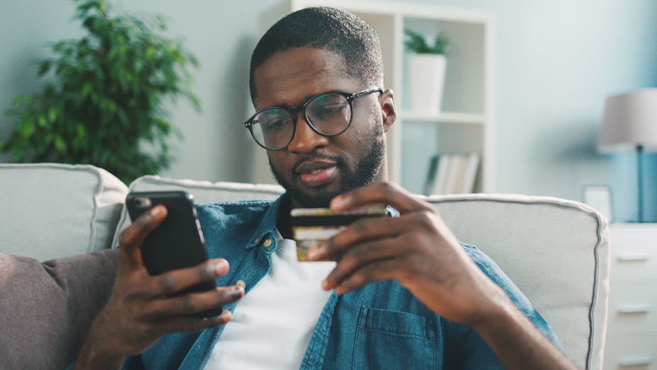 African young man in glasses shopping online with credit card using smart phone at home.