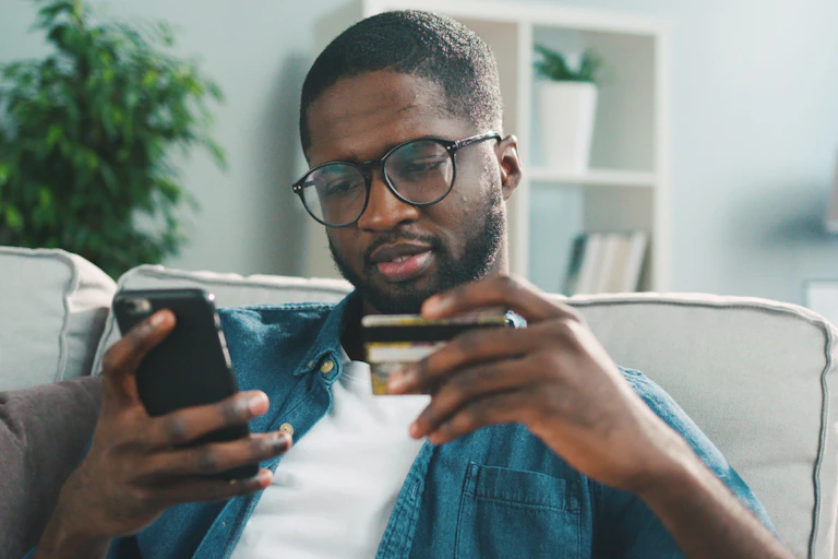 African young man in glasses shopping online with credit card using smart phone at home.