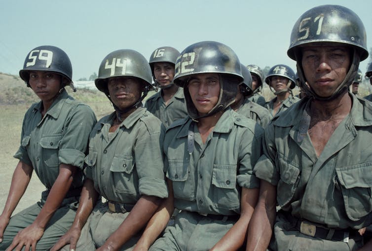 Half a dozen young men in green military gear and helmets sit nervously and look straight at the camera