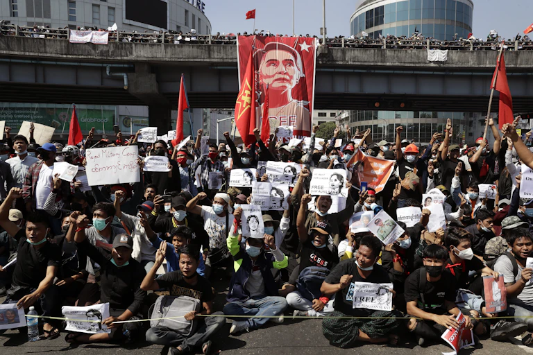 Protesters sitting on ground in front of banner with Aung San Suu Kyi