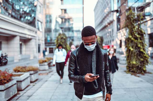 A man wearing a surgical mask walking down a city street looking at his phone