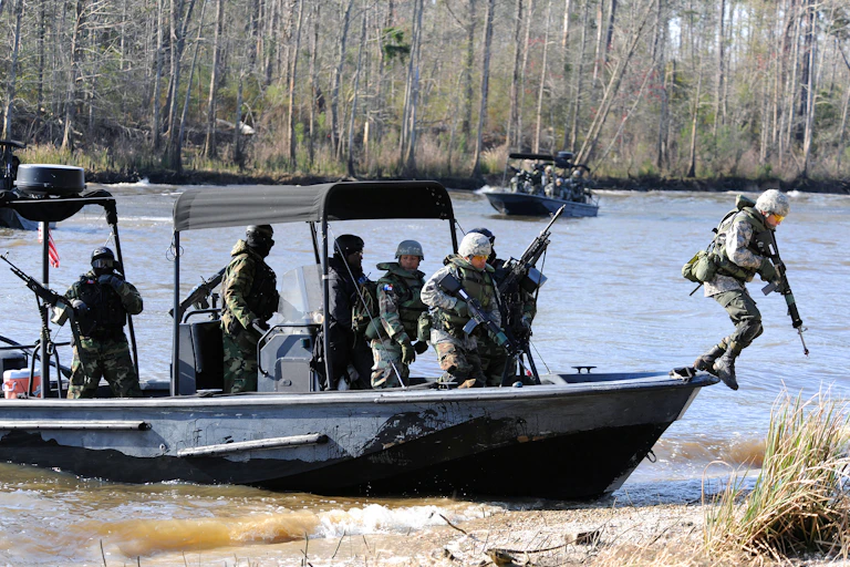 Seven soldiers in fatigues carrying weapons disembark from a small craft to a rocky shoreline