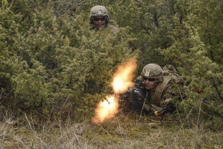 Photo of two soldiers in fatigues, one standing and the other lying in the bushes shooting a weapon