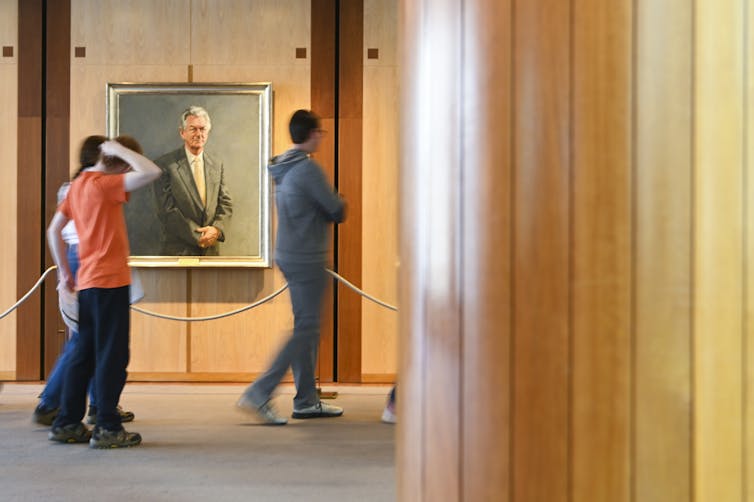 Three people walk past a portrait hanging in a wood-panelled room.