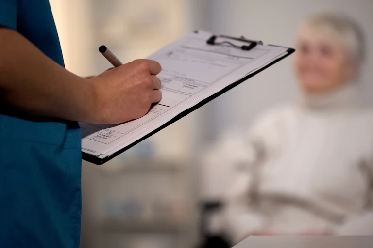 A health-car worker taking notes on a clipboard with a woman with a disability in the background