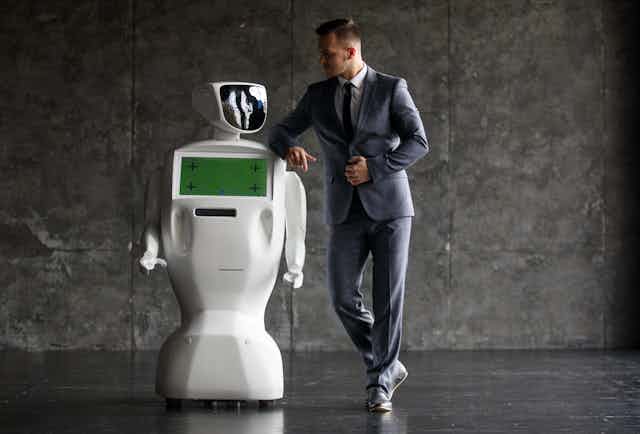 A man in a suit leans casually on the shoulder of a basic-looking robot