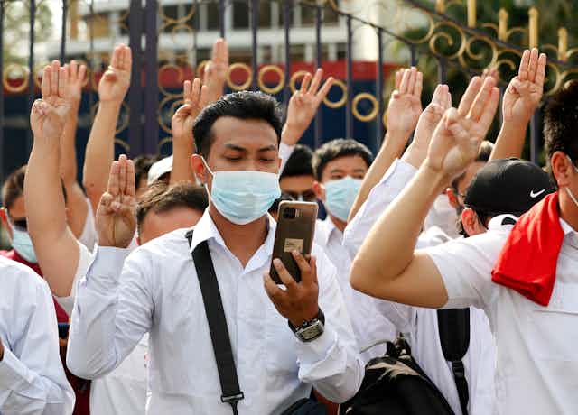 Group of young men and women in white shorts and COVID masks with their left hands using three-fingered protest salute from The Hunger Games.