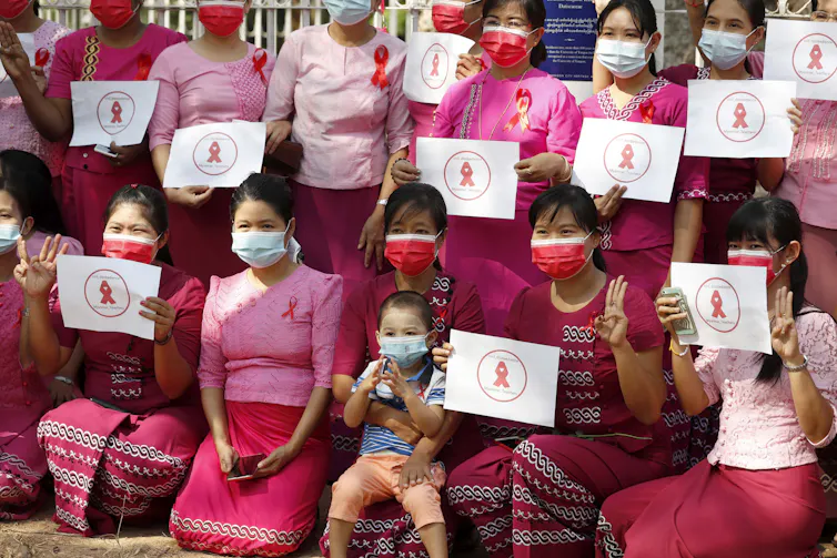 Young women wearing pink uniforms and COVID masks hold up placards with the red scarf resistance symbol.