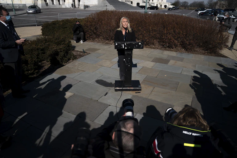 Greene speaks from a podium while a crowd of reporters and photographers surround her.