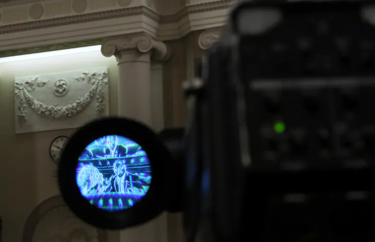 A congressman on the House floor is shown through the lens of a camera.