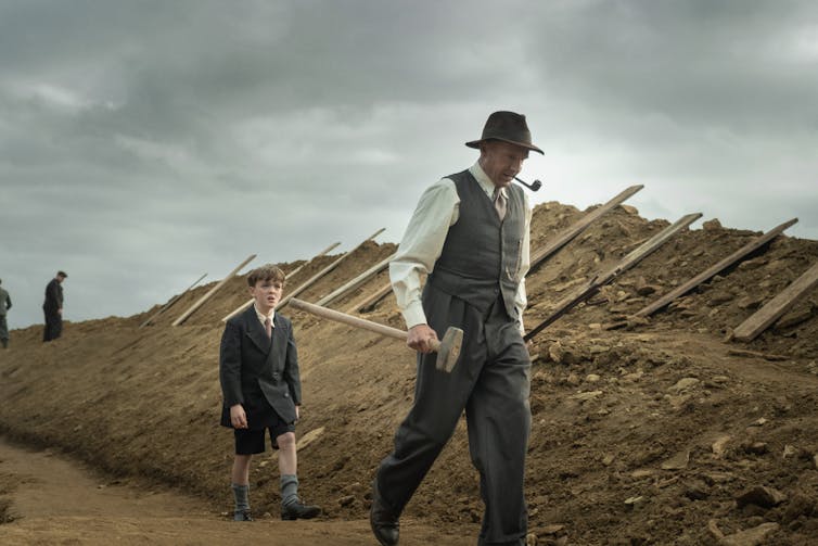 Film still. Basil holds a mallet and walks past a mound of dirt. He is followed by a young boy.