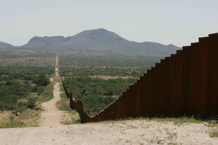 A border wall running along a dirt track in a hot, dry plain.