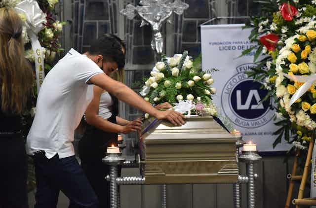 Man in white t-shirt leans on a coffin with flower displays at a church in Buga, Valle del Cauca, Colombia.