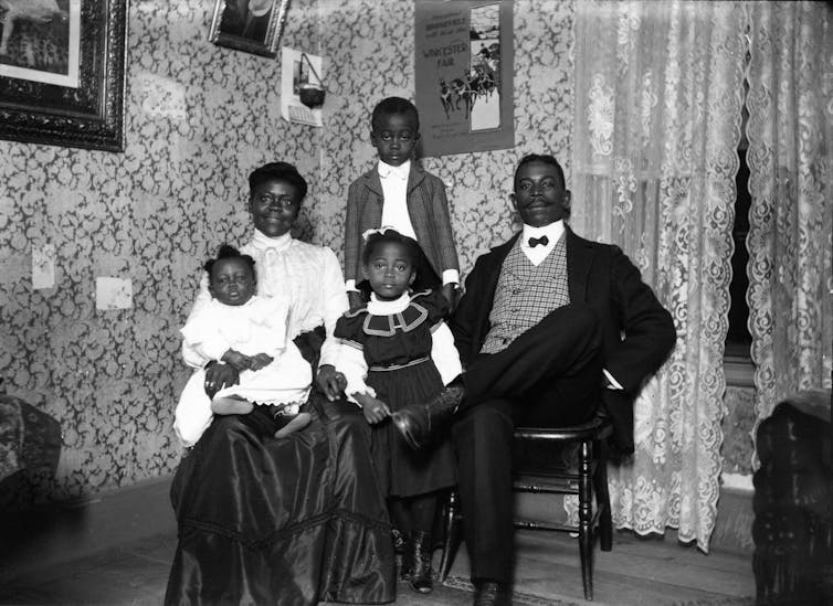 The family, proudly dressed, is photographed indoors, in front of lace curtains and framed prints.