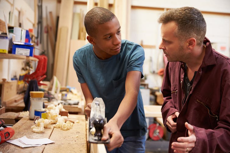Mentor talks to young male apprentice planing some wood
