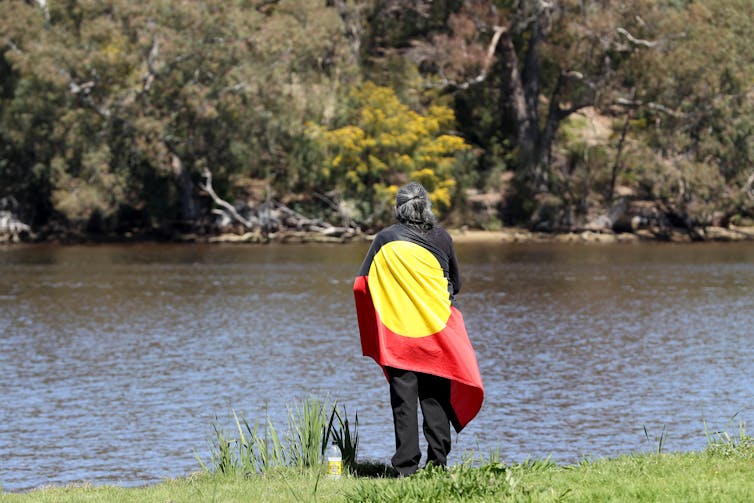 Man wrapped in Aboriginal flag stands on river bank.
