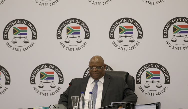 A man wearing specs, a dark suit and blue tie sits in a chair.