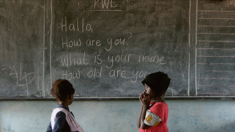 Two children in front of a blackboard showing English phrases.