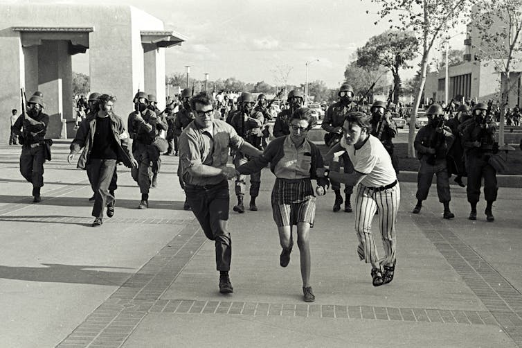 Black-and-white image of three running young people, chased by a dozen armed soldiers