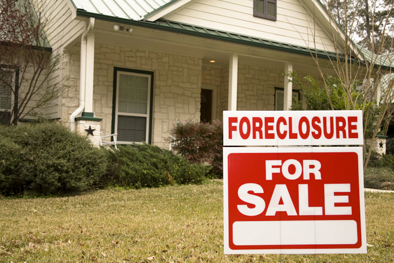 Photo of a white home with green roof and a "foreclosure" sign in the yard