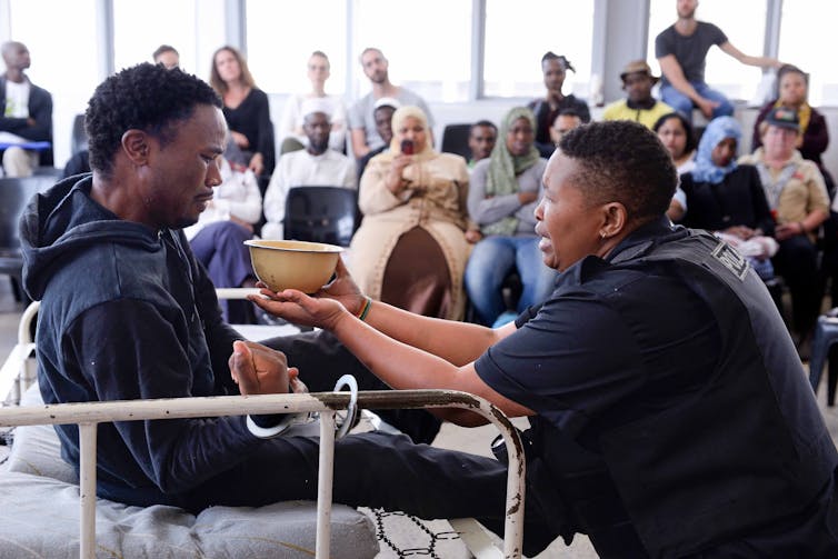 An audience watches a pained middle-aged woman hold a bowl towards the mouth of a man handcuffed to a bed.