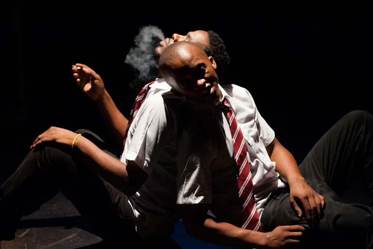 Lit up in the dark, two men sit, leaning their backs against one another, one exhaling a plume of smoke from a hand-rolled cigarette.