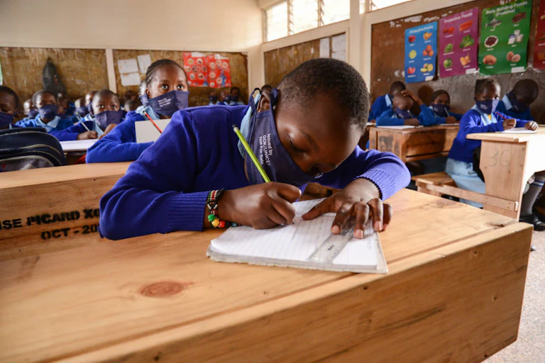 A classroom full of young learners, wearing face masks and blue uniforms they are focused on writing in the books on the desks in front of them.