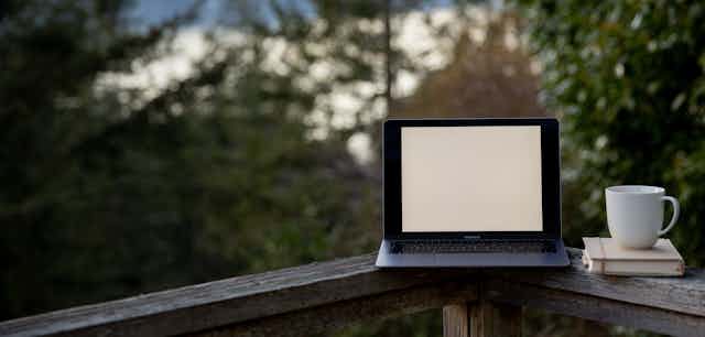 A laptop sits on the railing of a deck overlooking a lake, trees and hills.