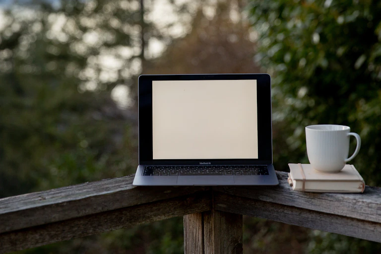 A laptop sits on the railing of a deck overlooking a lake, trees and hills.