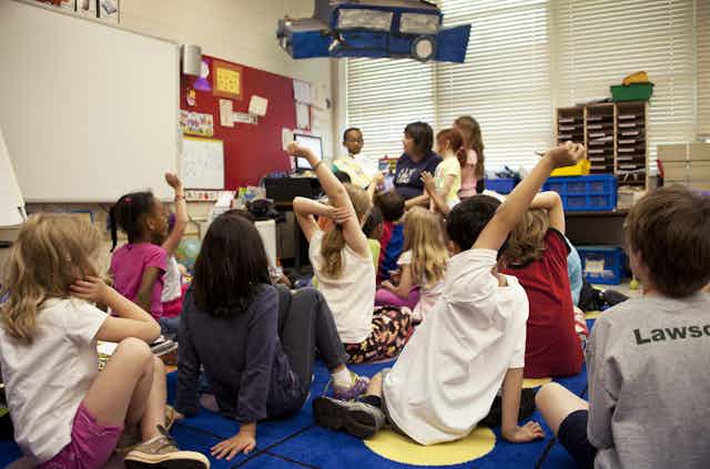 Kids raise their hands in a middle-school classroom. Children look about 7-years old. 