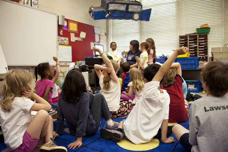 Kids raise their hands in a middle-school classroom. Children look about 7-years old.