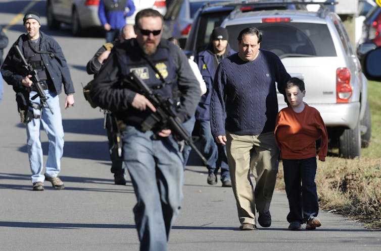 A parent with a child walks near police officers with rifles.