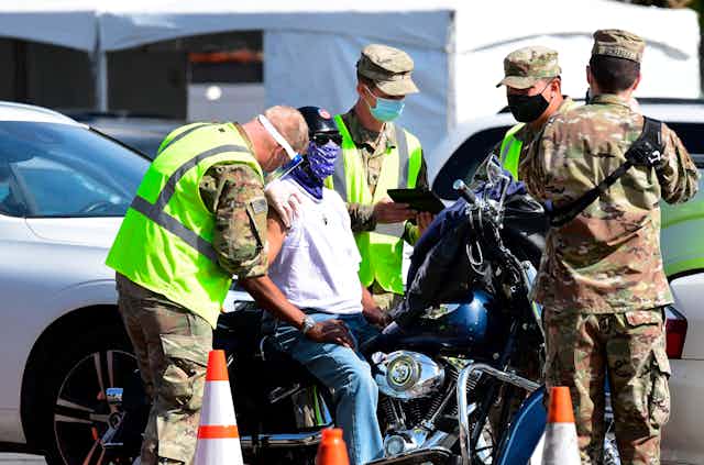 A man on motorcycle gets a vaccine shot from National Guard troops.