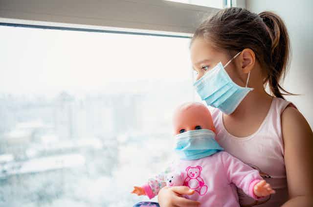 Girl wearing mask looking out of window holding doll wearing mask