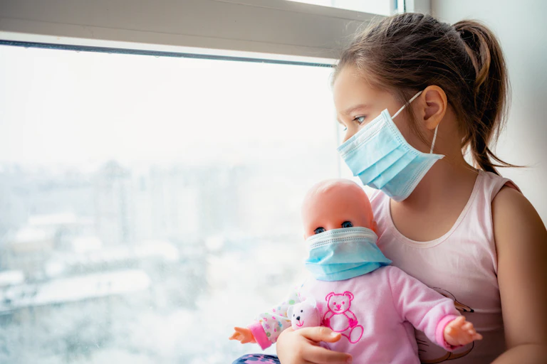 Girl wearing mask looking out of window holding doll wearing mask
