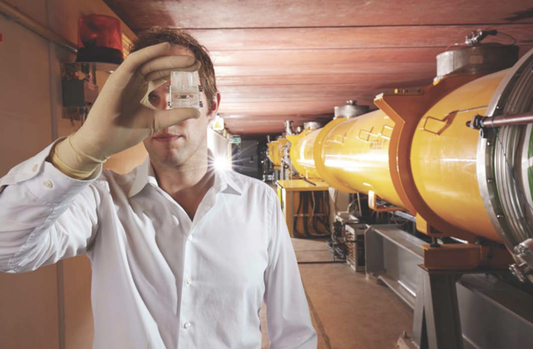 A man holds a clear component in front of his eye. Behind him is a large yellow pipe
