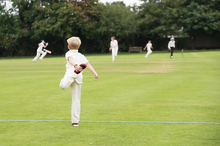 A boy dressed for playing cricket stretches his leg, with other players in the background.