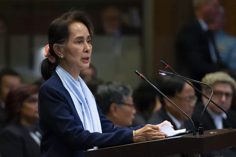 Aung San Suu Kyi stands before a podium and speaks.