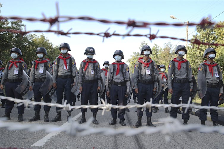 A row of police in riot gear stand behind barbed wire.