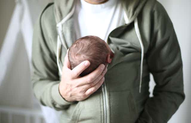 A man's right hand holds the head of a baby who is held inside his jacket. 