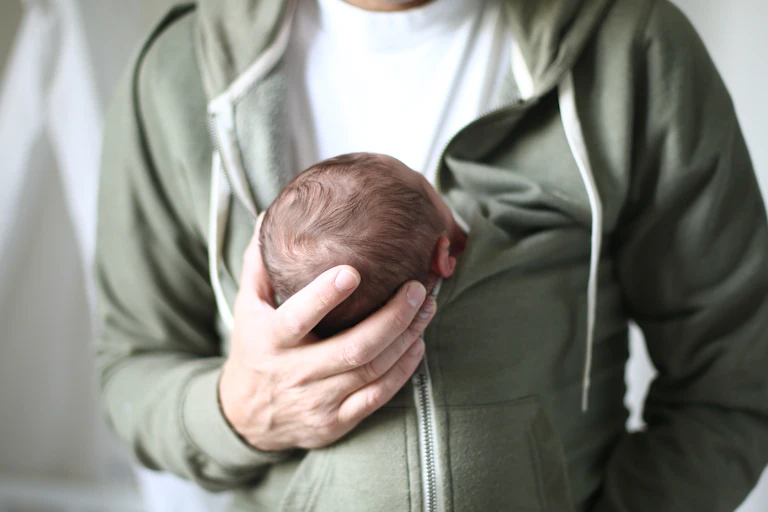 A man's right hand holds the head of a baby who is held inside his jacket.