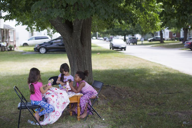 Three young girls play with dolls together at a table outside.