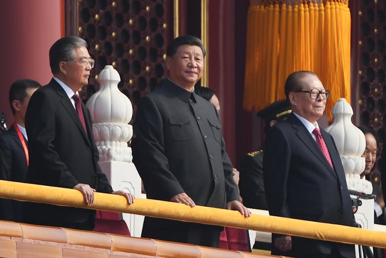 Xi stands on a balcony in black suit with a Mao-style collar, flanked by his predecessors