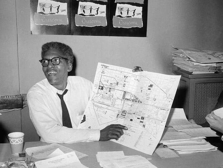 Black-and-white image of Rustin at a desk holding a big map and smiling, with papers all over this desk