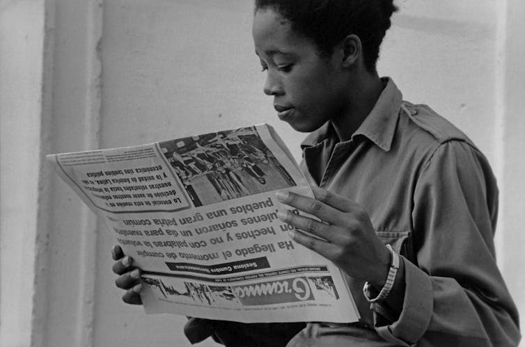 Black-and-white image of a young woman in fatigues reading the paper