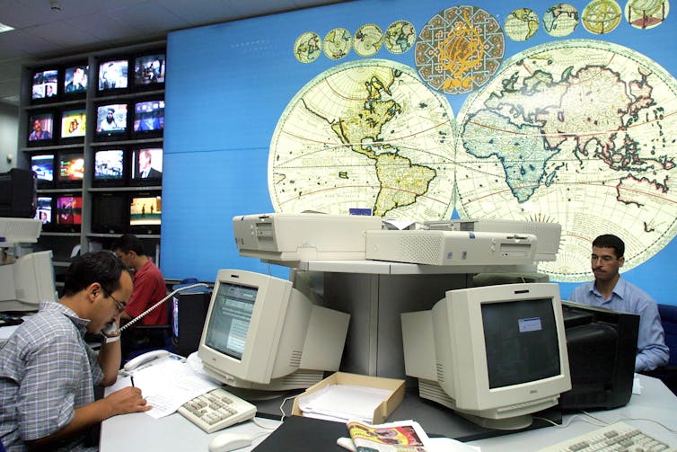 Two journalists sit in a newsroom with old desktop computers and a world map on the wall