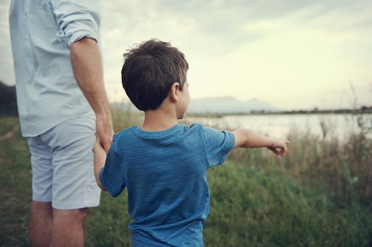 Young boy pointing to something, while holding his fathers hand. They're standing in front of a lake.