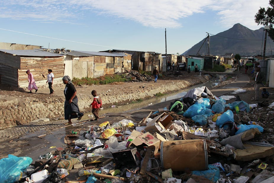 Woman and child walk past piles of rubbish next to an open drain and a row of shacks